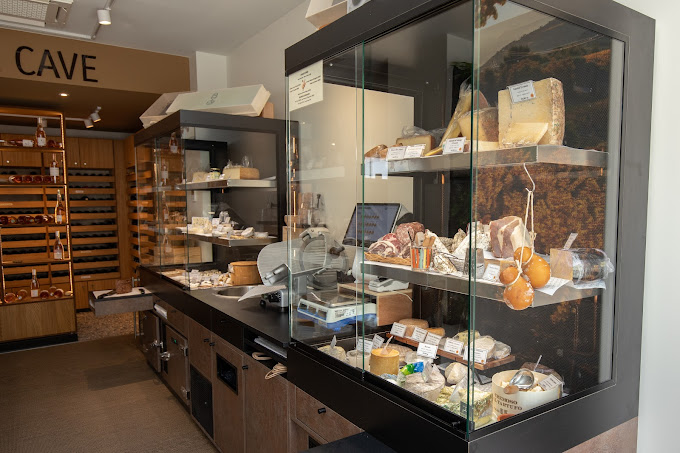Une vitrine réfrigérée remplie d'une variété de fromages et de charcuteries, ainsi que d'autres produits frais. Sur la gauche, des étagères en bois sont remplies de bouteilles de vin. Le mot "CAVE" est visible en haut à gauche.