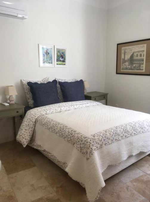 Une chambre lumineuse et accueillante avec un lit double orné d'une couette blanche à motifs floraux et de grands oreillers bleus. Deux tables de nuit grises avec des lampes flanquent le lit, sous des œuvres d'art encadrées, et un sol carrelé beige.