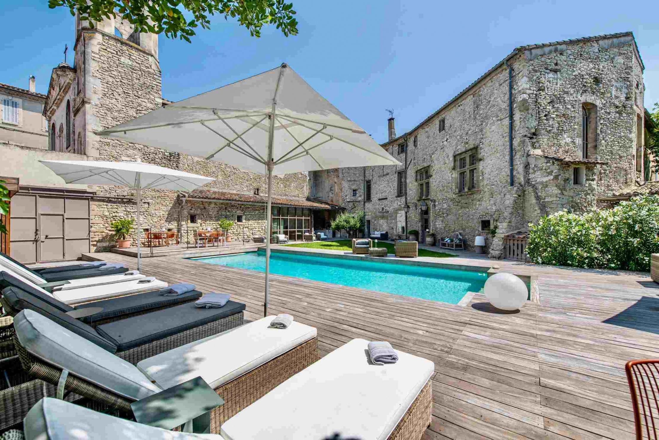 Piscine extérieure avec terrasse en bois, chaises longues et parasols, entourée de bâtiments en pierre.