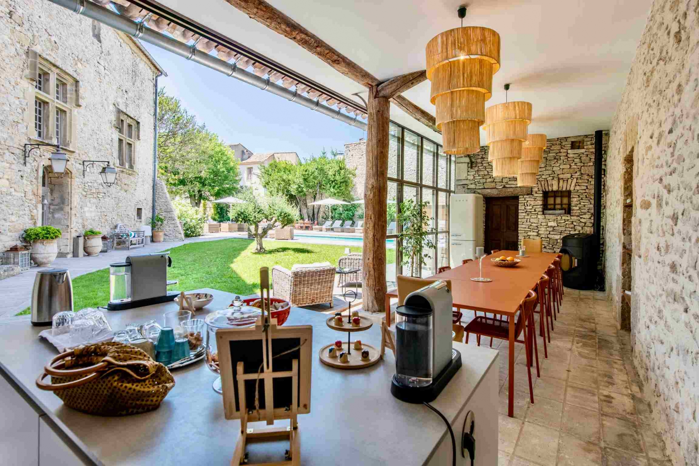 Cuisine moderne avec comptoir, machines à café, longue table à manger, lustres, et vue sur un jardin verdoyant et une piscine.