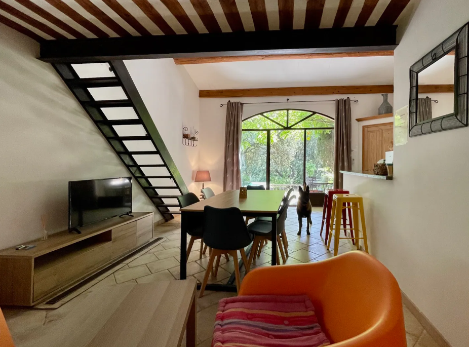 Salon spacieux avec un escalier moderne, une télévision, une table à manger, un bar et un chien près d'une grande fenêtre.