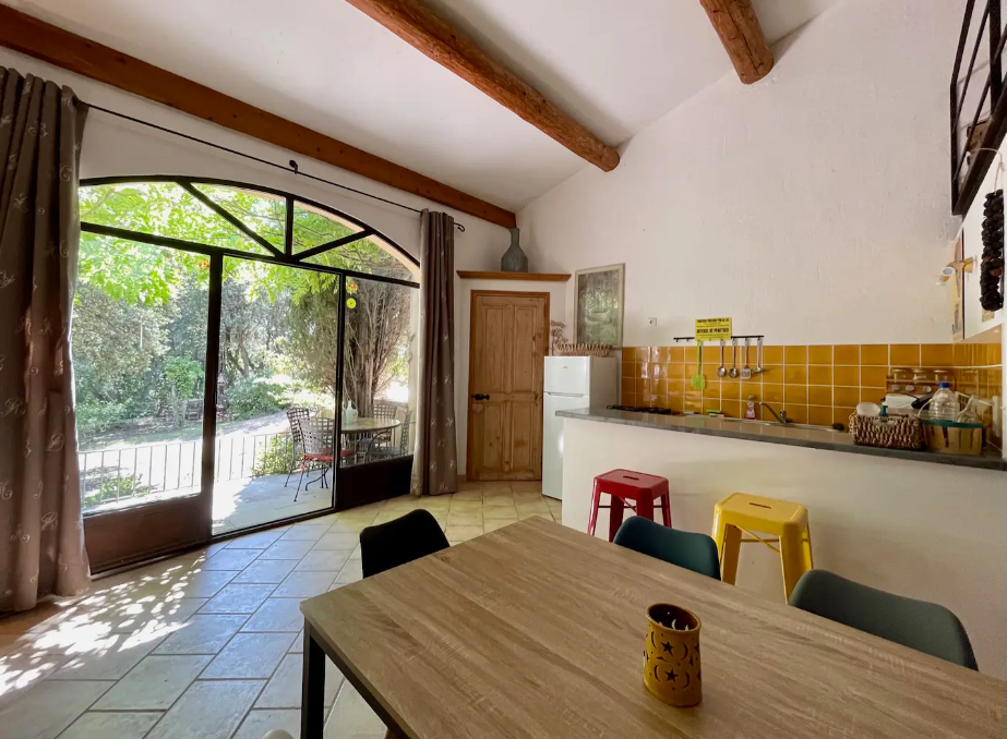 Cuisine rustique avec poutres apparentes, table en bois, tabourets colorés et grande porte vitrée donnant sur un jardin verdoyant.
