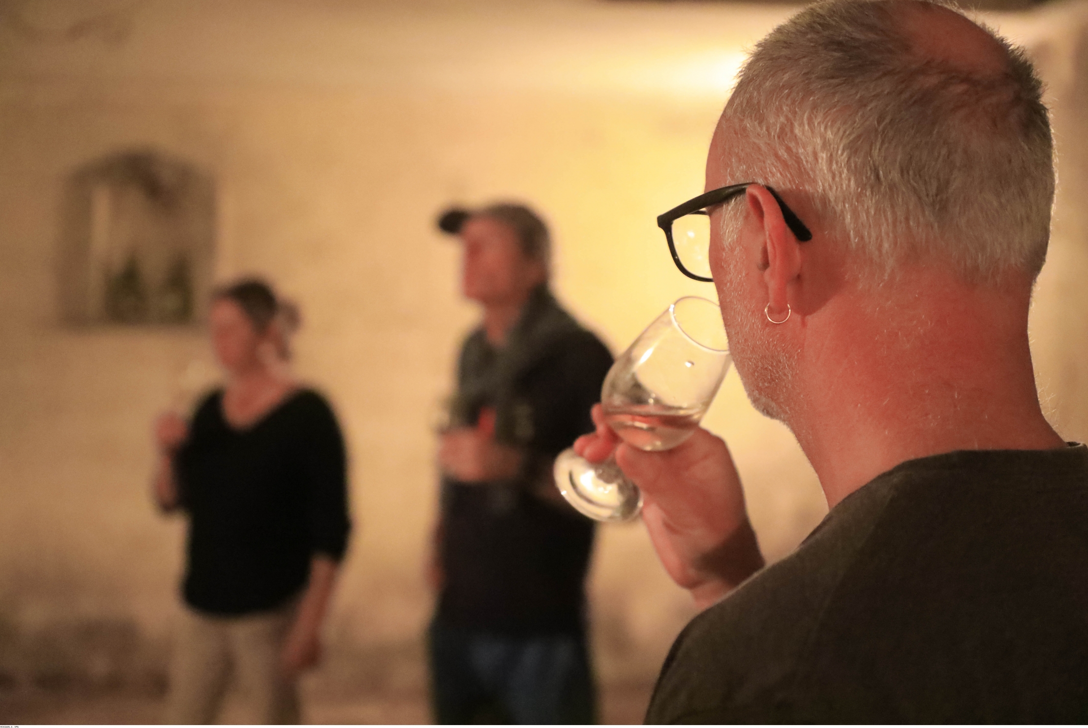 Homme aux cheveux gris et lunettes noires tenant un verre de vin dans un cadre éclairé.