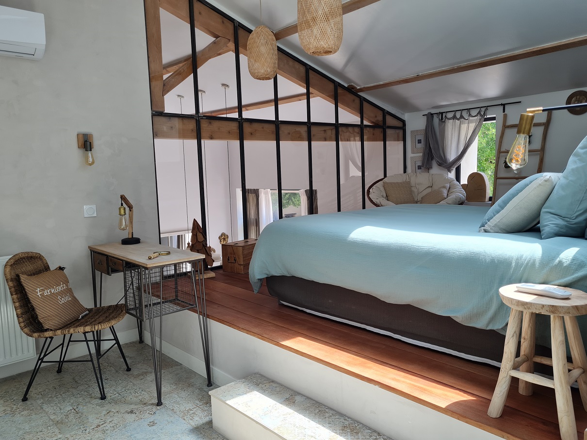 Une chambre spacieuse et moderne, avec un lit bleu turquoise sur une estrade en bois, et un espace bureau séparé par une cloison vitrée.