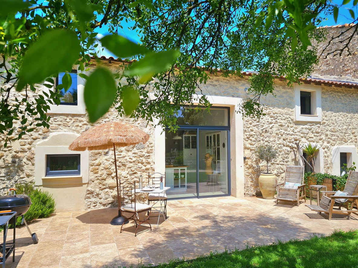 Une mas provençal en pierre avec une terrasse ensoleillée, un parasol, des meubles de jardin en bois et un barbecue, entouré de verdure. Idéal pour des vacances relaxantes.