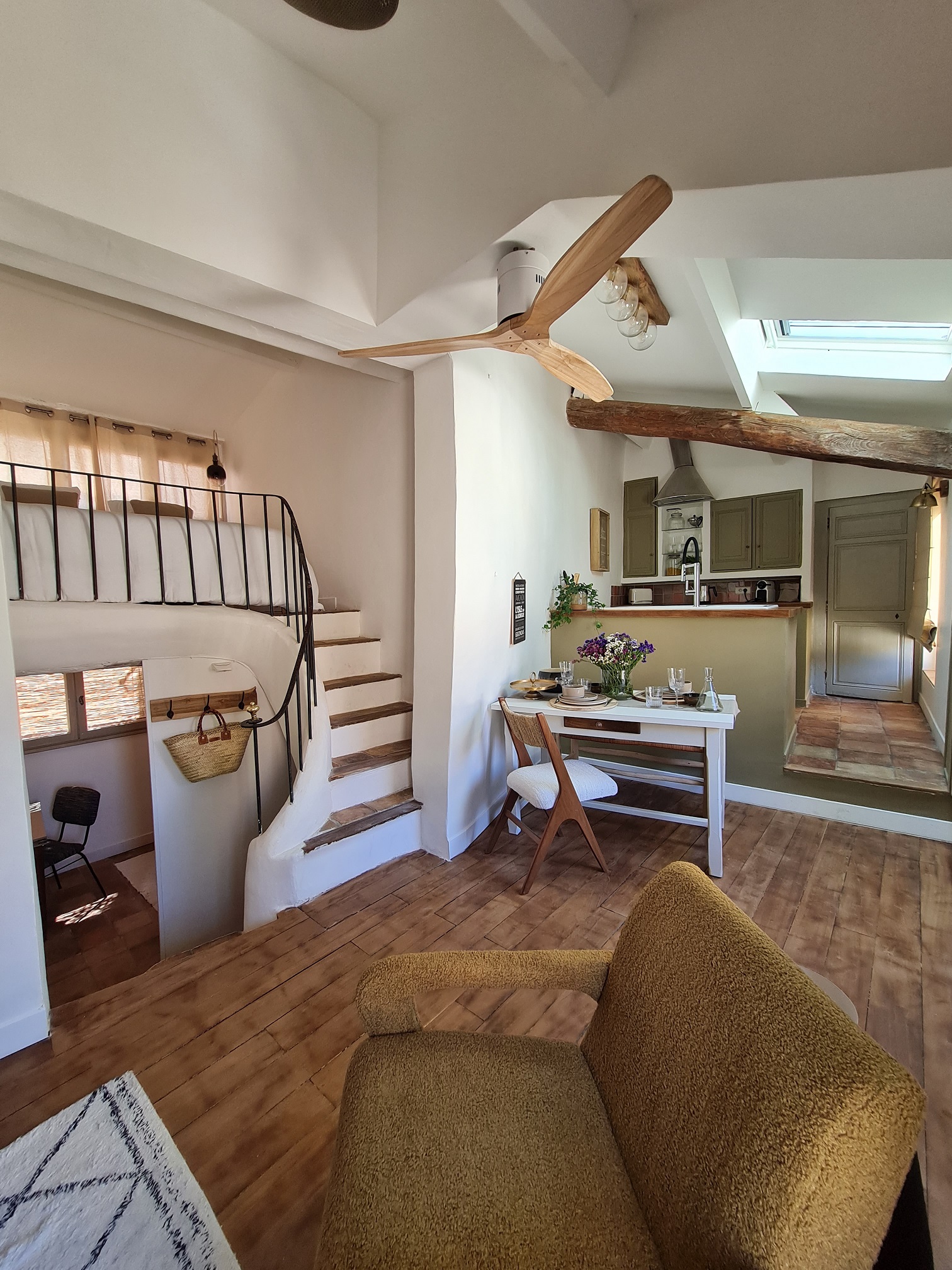 Une pièce à vivre lumineuse avec un coin cuisine, une table à manger dressée, un salon avec un fauteuil moelleux, et un escalier menant à une mezzanine.