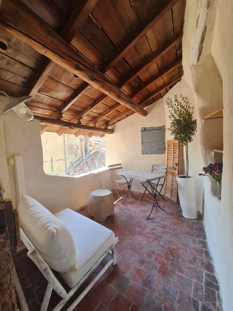 Une petite terrasse rustique avec des murs en pierre et un plafond en bois, meublée d'une table et de chaises en fer forgé, ainsi qu'un canapé blanc et un olivier en pot.