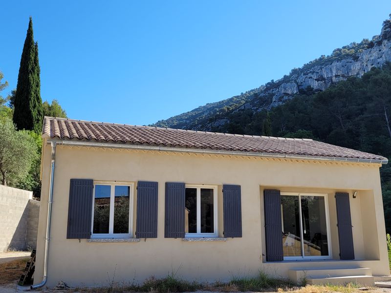 Une charmante maison provençale aux volets gris et au toit de tuiles, nichée au pied d'une imposante montagne verdoyante, avec un grand cyprès sur la gauche et un ciel bleu éclatant.