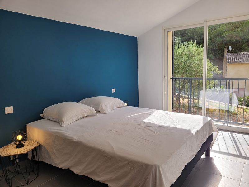 Une chambre lumineuse avec un grand lit double et un mur d'un bleu profond, ouverte sur un balcon et la nature environnante, avec une petite table de chevet et une lampe design.