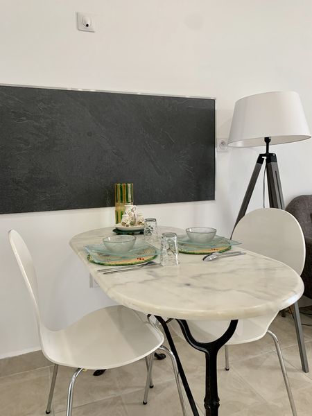 Une table à manger ovale en marbre blanc, avec deux chaises blanches, dressée pour deux personnes, sous un grand tableau noir, avec une lampe sur pied moderne en arrière-plan.