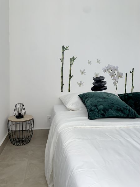 Une chambre minimaliste avec un lit blanc et des coussins verts, décorée de stickers muraux représentant des bambous, des fleurs d'orchidée et des pierres zen, avec une table de chevet moderne.