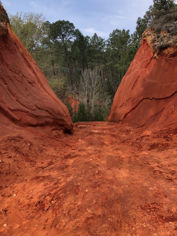 Une allée d'un sentier d'ocre en Provence, bordée de falaises rocheuses rouge, sous un ciel bleu clair.