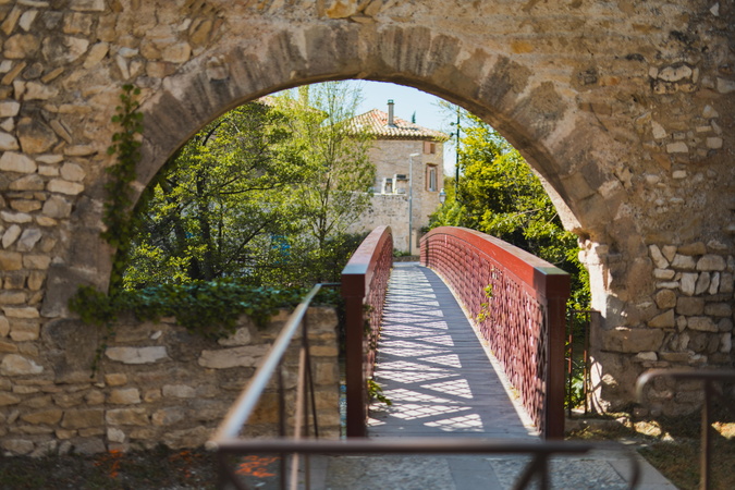 Un pont en bois avec des rambardes rouges s'étend à travers une arche en pierre, menant vers une maison et des arbres.