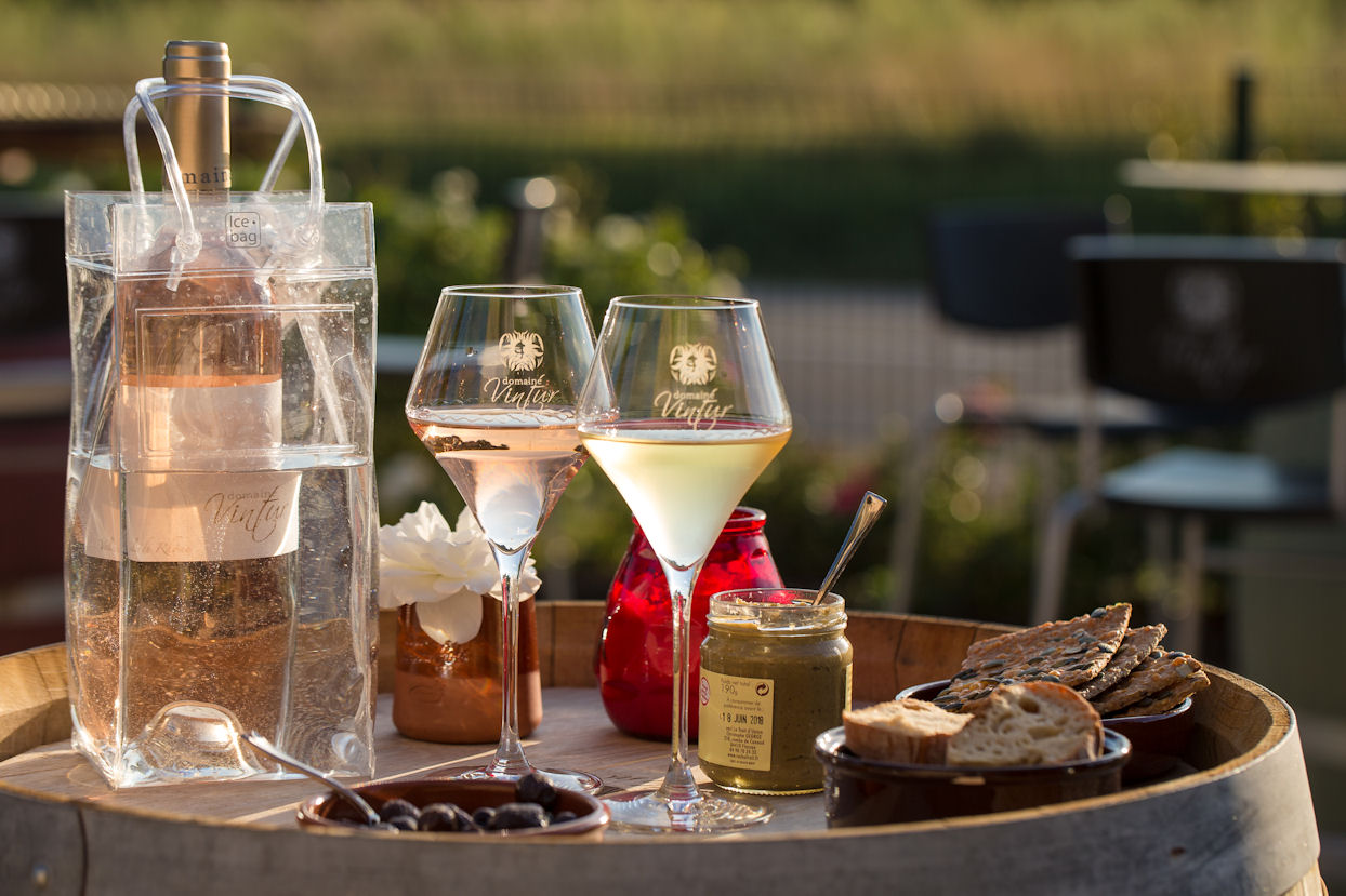 Une scène de dégustation en extérieur avec une bouteille de rosé dans un rafraîchisseur en acrylique, deux verres de vin (un de rosé, un de blanc), des olives, du pain, et des tartinades sur un tonneau en bois. L'arrière-plan flou suggère un vignoble.