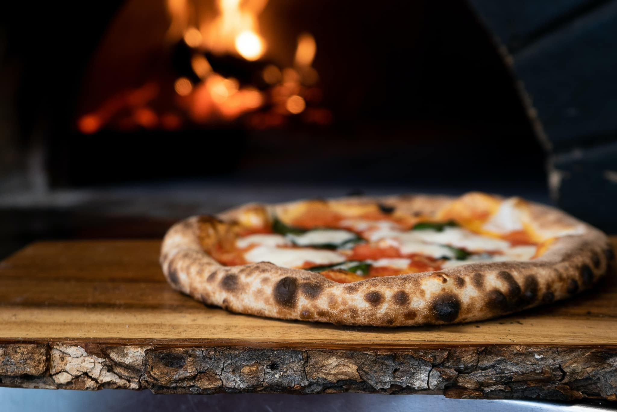 Une pizza fraîchement cuite avec une croûte croustillante, des tomates, de la mozzarella et du basilic, posée sur une planche en bois devant un four à bois.