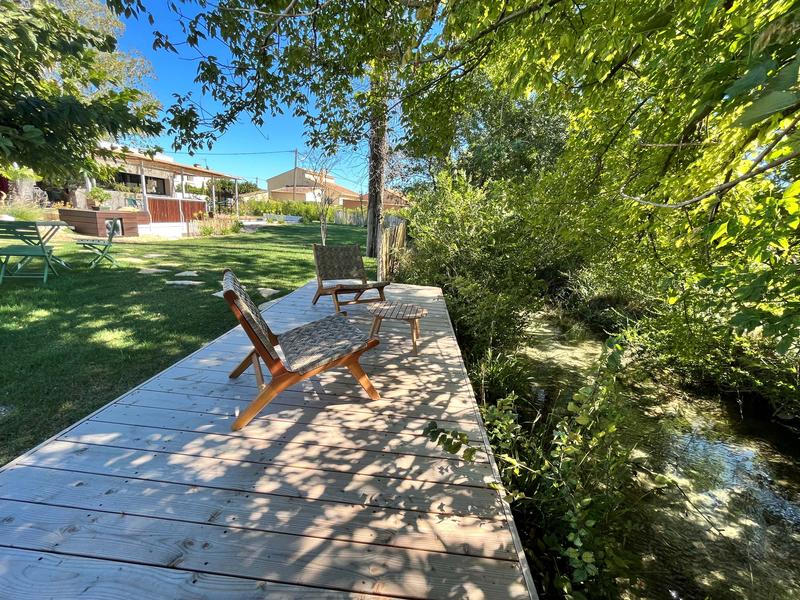 Une terrasse en bois avec des chaises longues, bordant un petit cours d'eau sous un grand arbre et un ciel bleu clair.