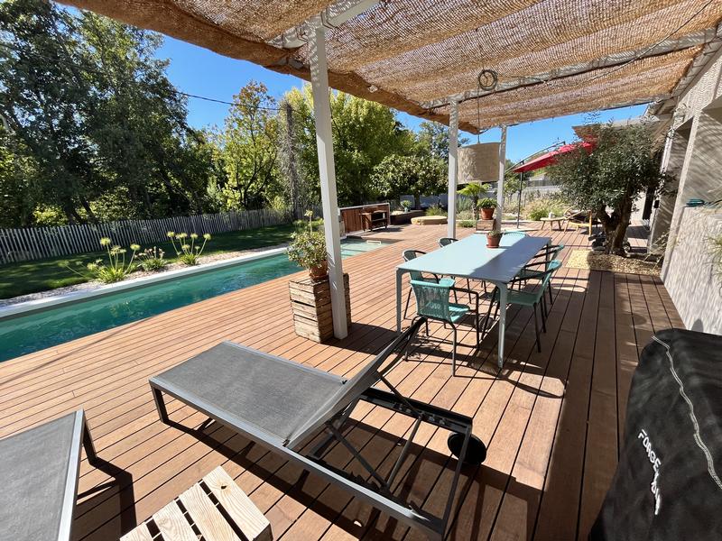 Une terrasse extérieure en bois avec une piscine, un salon de jardin, des chaises longues et un parasol, sous un ciel bleu.