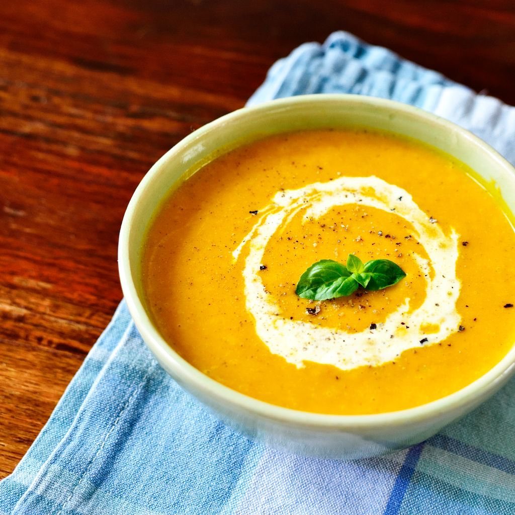 Un bol de soupe orange crémeuse garni d'une feuille de basilic et de poivre moulu, posé sur une serviette bleue à carreaux.