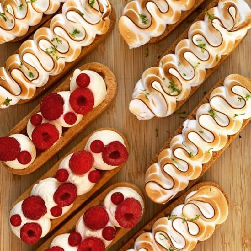 Des pâtisseries allongées, certaines avec une meringue dorée et du zeste vert, et d'autres garnies de crème blanche et de framboises fraîches.