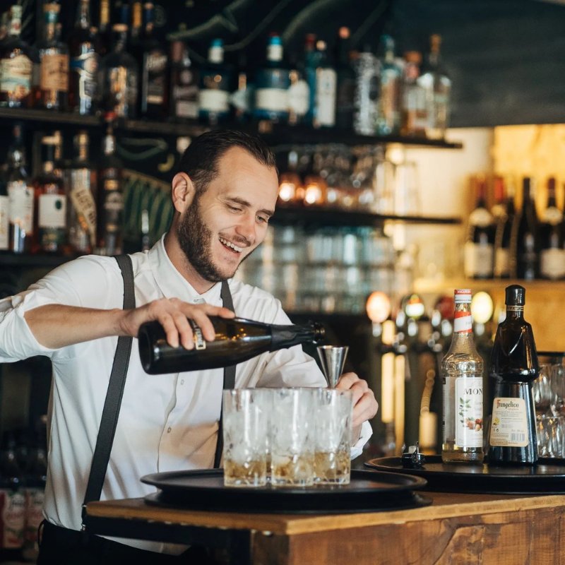 Un barman souriant en bretelles verse une boisson d'une bouteille dans un verre doseur, avec des bouteilles d'alcool et des verres en arrière-plan.