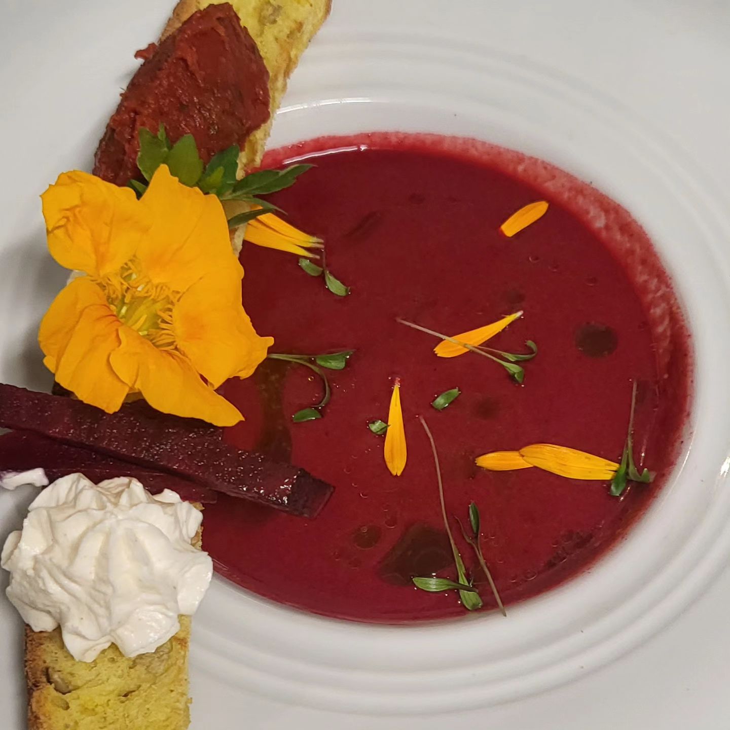 Un bol de soupe rouge garni de pétales de fleurs jaunes, de fines herbes et d'une quenelle de crème.