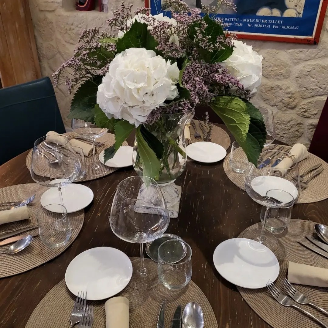 Une table ronde de restaurant dressée avec des verres, des assiettes blanches, des couverts et un grand bouquet de fleurs blanches et violettes au centre.