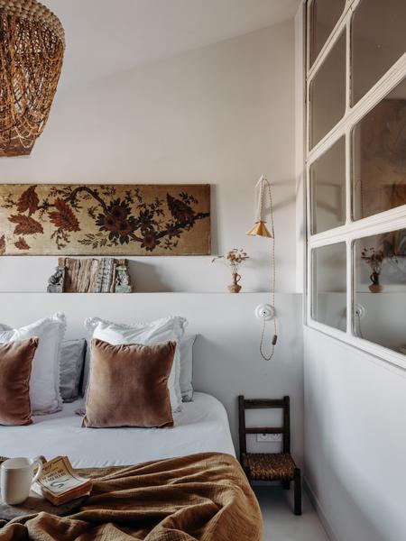 Une chambre lumineuse avec un lit fait, un tableau mural floral, une suspension en osier, une lampe et une petite chaise en bois.