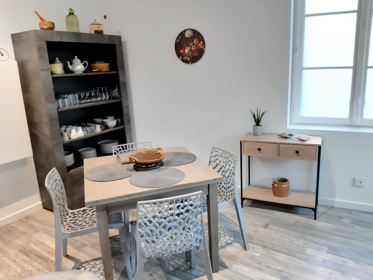 Une salle à manger moderne avec une table et quatre chaises, une étagère remplie de vaisselle et une commode en bois à côté d'une fenêtre.