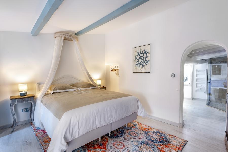 Une chambre lumineuse avec un lit double orné d'un baldaquin et d'une literie beige et blanche, reposant sur un tapis coloré aux motifs vifs. Des poutres bleues traversent le plafond blanc, et des tables de chevet avec des lampes flanquent le lit. Une salle de bain est visible à travers une arche.