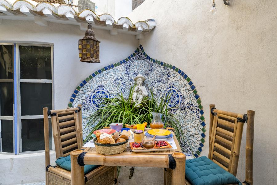 Un petit-déjeuner coloré est dressé sur une table en bambou avec deux chaises, devant un mur orné d'une mosaïque bleue et blanche et d'une statue. Une lanterne est suspendue au-dessus.