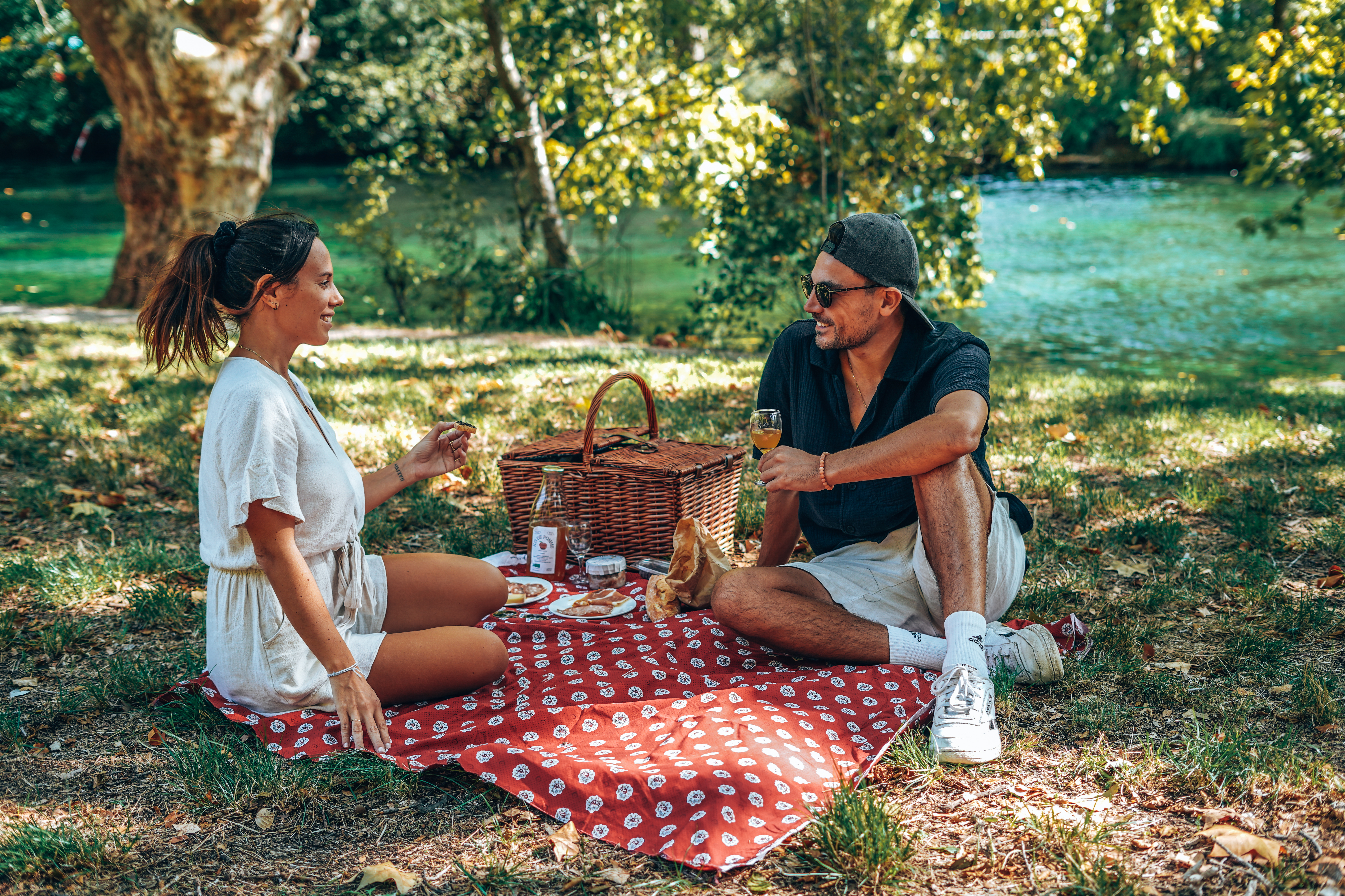 Un couple est assis sur une couverture de pique-nique à carreaux rouges et blancs dans un parc, sous un grand arbre.