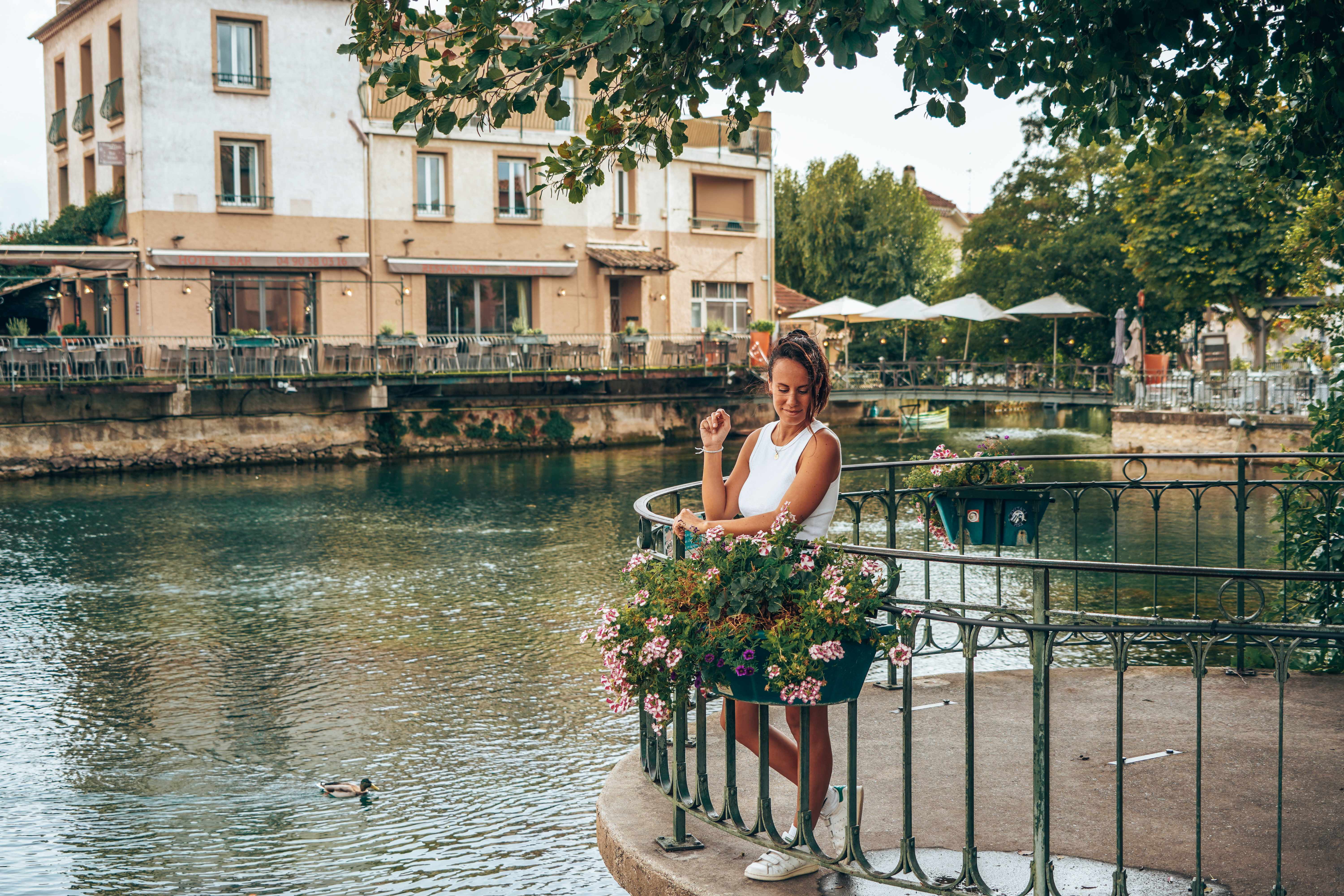 Une jeune femme souriante, appuyée sur une balustrade fleurie au bord d'une rivière, avec des bâtiments en arrière-plan.