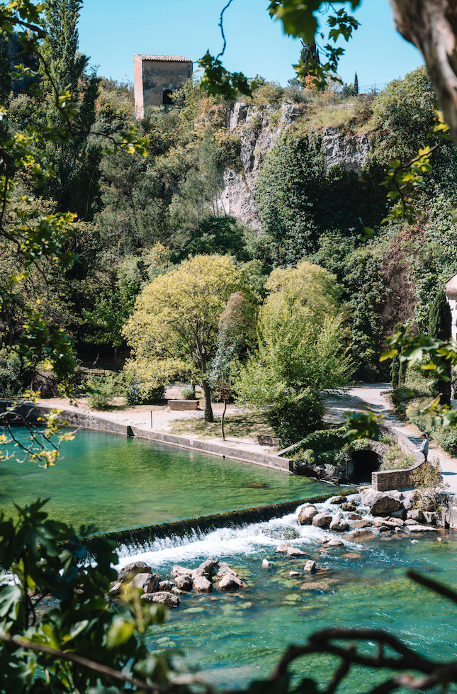 Une cascade coule dans une rivière verte, entourée d'arbres verdoyants, avec un bâtiment en pierre sur la colline en arrière-plan.