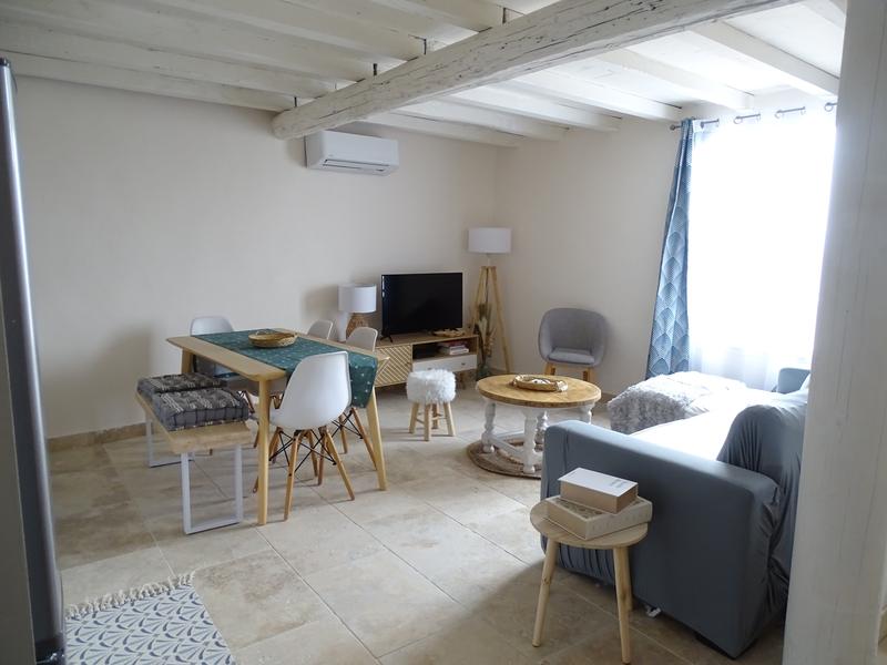 Un salon ouvert avec une table à manger en bois, des chaises blanches, et un canapé gris devant une télévision.