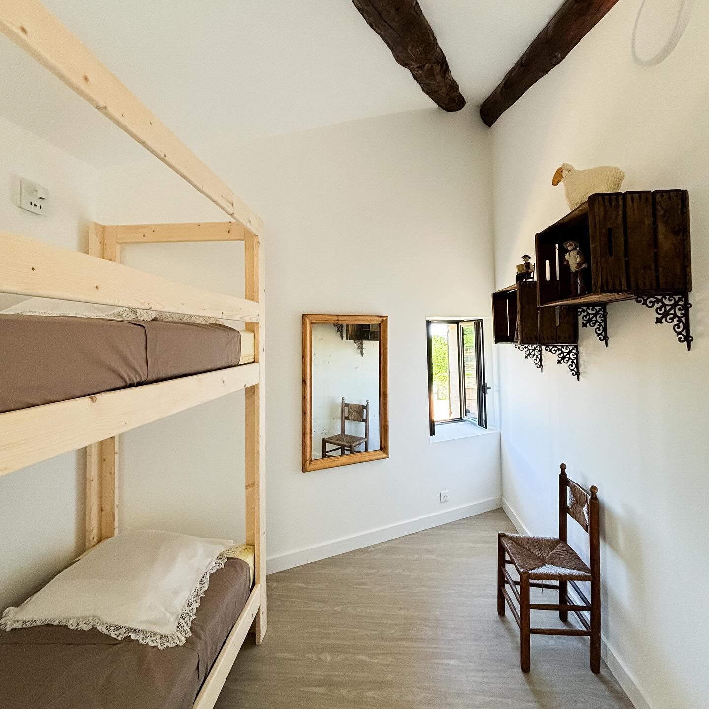 Une chambre lumineuse avec un lit superposé en bois, un miroir mural, une petite fenêtre ouverte et des étagères décoratives en bois.