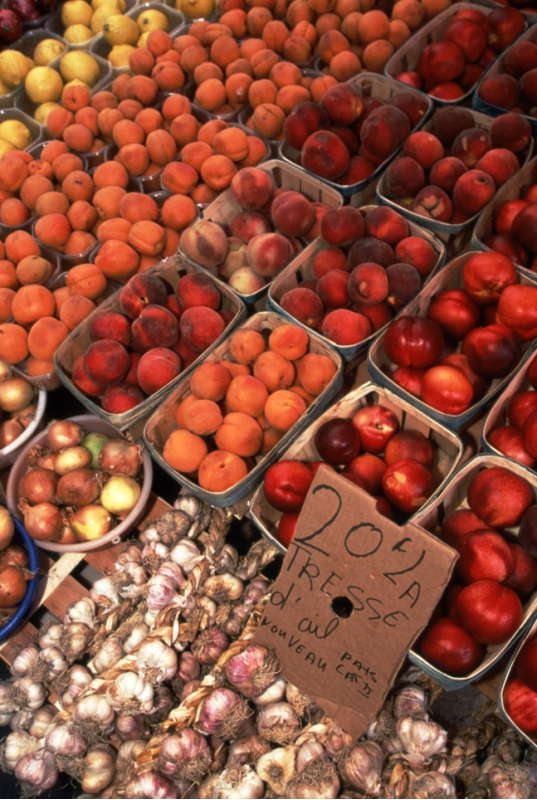 Un étal de marché coloré regorge de cagettes de fruits frais, principalement des pêches et des abricots. Au premier plan, de l'ail frais tressé est exposé avec une pancarte indiquant son prix et sa provenance.