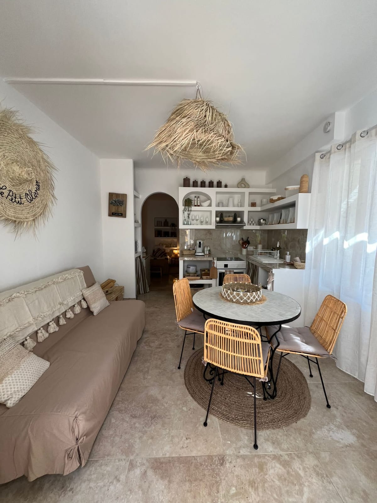 Un intérieur lumineux avec un canapé beige, une table ronde avec des chaises en rotin et une cuisine ouverte avec des étagères.
