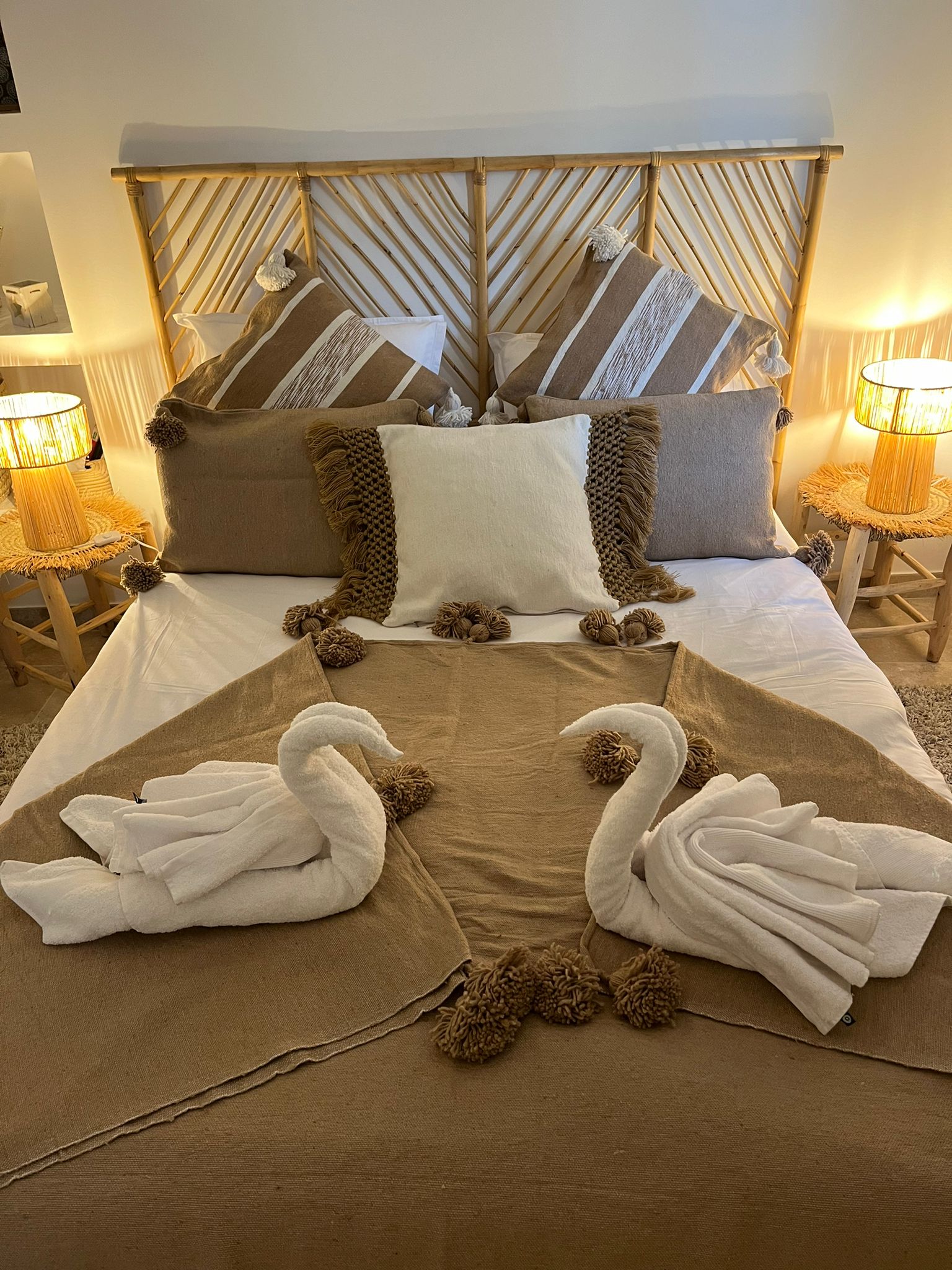 Une chambre douillette avec un lit fait de draps blancs et d'un plaid marron, décoré de coussins à pompons et de deux cygnes en serviettes, avec une tête de lit en bois et des lampes de chevet.