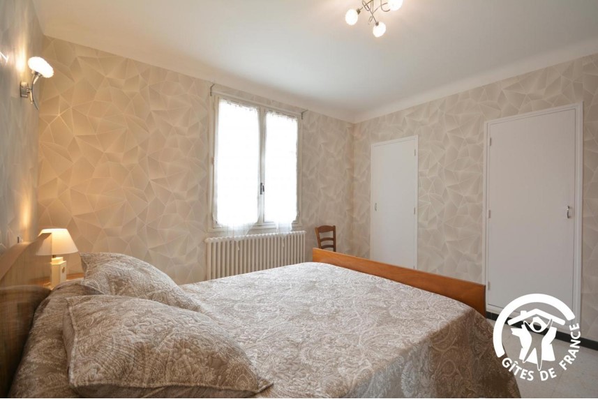 Une chambre lumineuse avec un grand lit double, des murs au motif texturé beige, une fenêtre avec un store et un radiateur en dessous, et deux portes blanches.