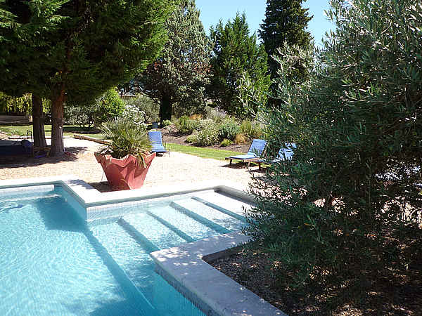 Une piscine aux marches immergées, bordée d'un aménagement paysager méditerranéen avec des arbres, des arbustes, un grand pot de fleurs et des chaises longues sous un soleil éclatant.