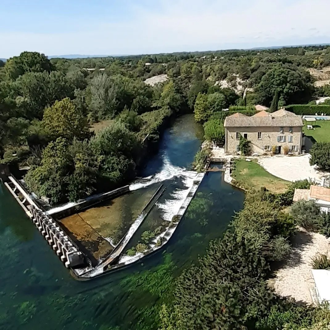 Une vue aérienne d'une rivière serpentant à travers un paysage verdoyant, avec un bâtiment en pierre et un barrage qui crée une cascade, sous un ciel clair.