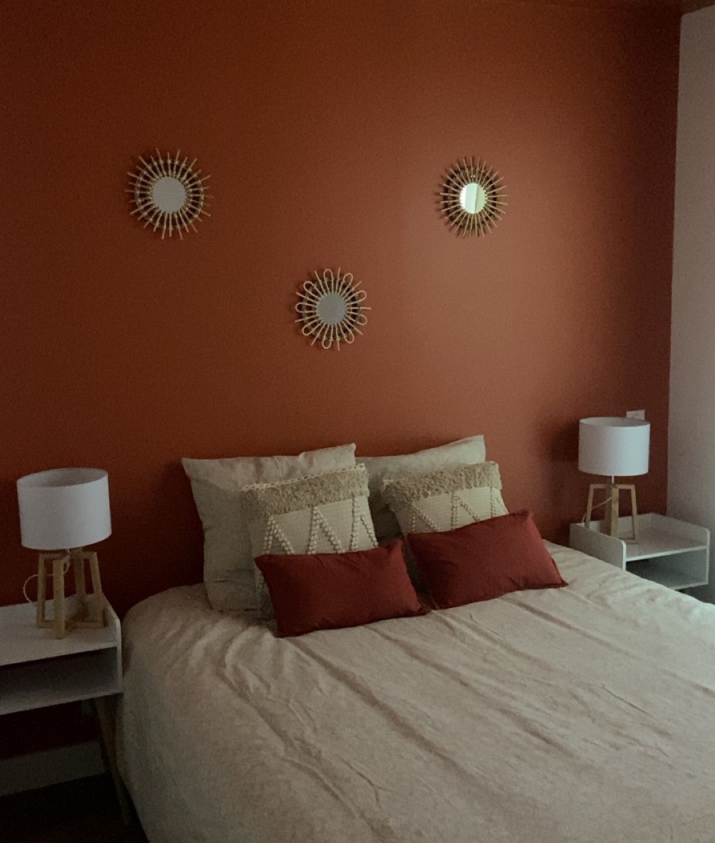 Une chambre à coucher aux murs terracotta, avec un grand lit beige orné de coussins rouges et blancs, flanqué de deux tables de chevet blanches surmontées de lampes.