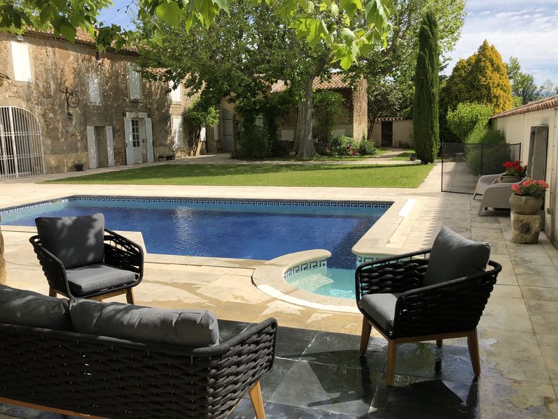 Une cour extérieure aménagée avec une piscine aux bordures claires, entourée d'une pelouse, d'arbres et d'une vieille bâtisse en pierre, avec un ensemble de mobilier de jardin moderne.