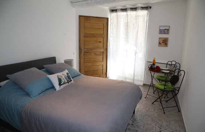 Une chambre simple et lumineuse avec un grand lit double recouvert d'une couette grise et de coussins bleus, une porte en bois clair, et un petit coin repas avec une table ronde en fer forgé et deux chaises à côté d'une fenêtre.