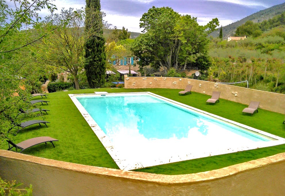 Une grande piscine rectangulaire entourée d'une pelouse verte et de chaises longues, avec des arbres, un cyprès et une maison de campagne en arrière-plan, sous un ciel nuageux.