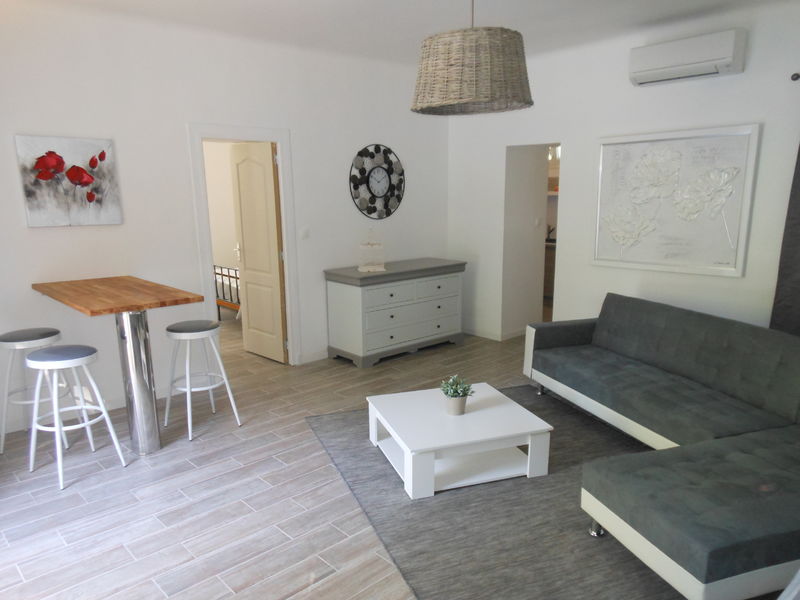 Un salon moderne et lumineux avec un canapé d'angle gris, une table basse blanche, un bar avec des tabourets hauts, une commode grise et blanche, et des œuvres d'art murales, sous une suspension en osier.
