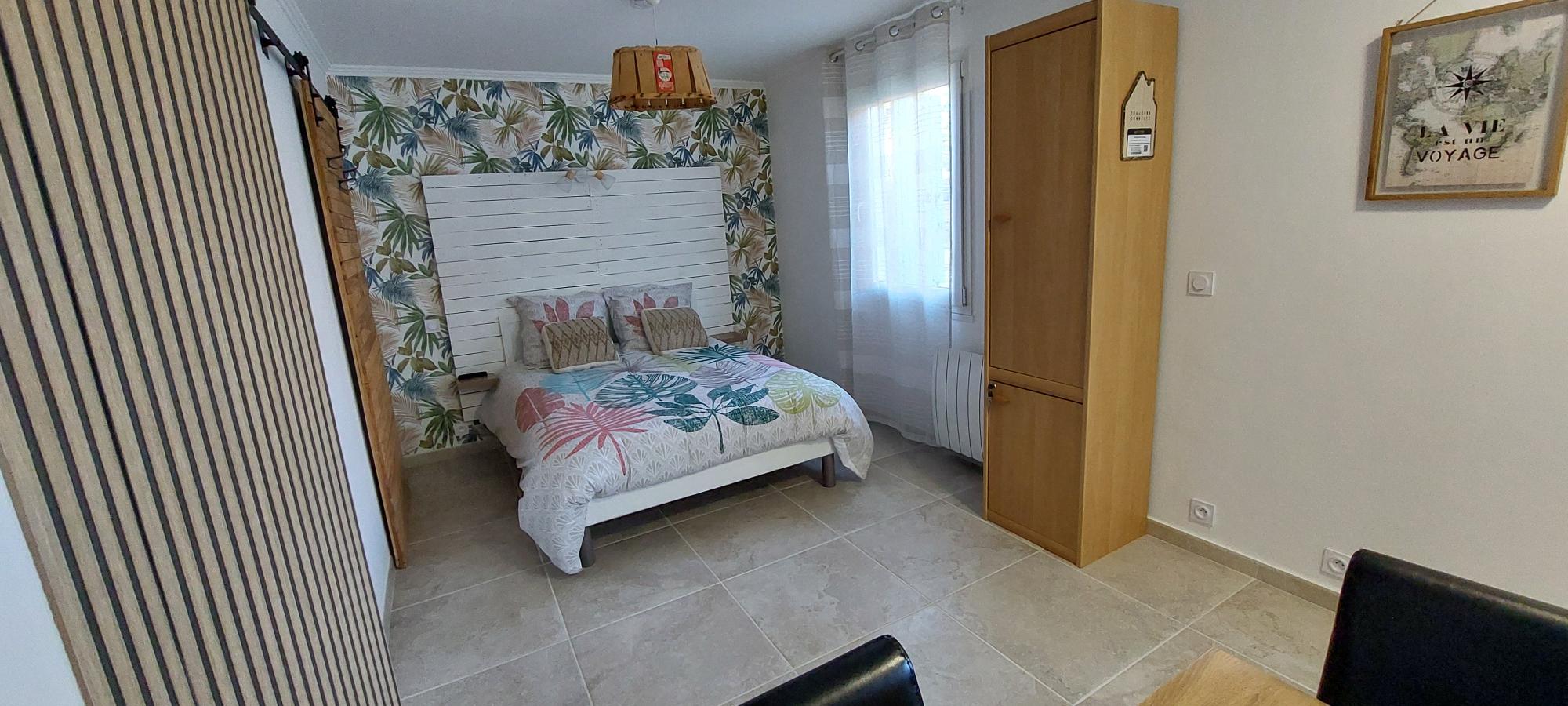 Une chambre lumineuse avec un lit double orné d'une couette à motifs tropicaux, une tête de lit en bois blanc, un mur tapissé de feuilles, une armoire en bois clair, et une fenêtre avec des rideaux blancs.