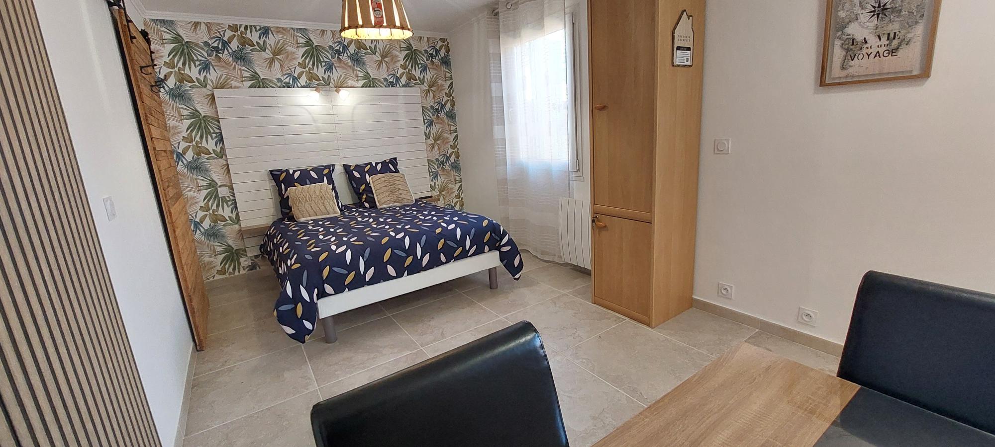 Une chambre moderne avec un lit double recouvert d'une couette bleue à motifs feuilles, une tête de lit en bois blanc, et un mur tapissé de motifs tropicaux, ainsi qu'une armoire en bois et un coin table.