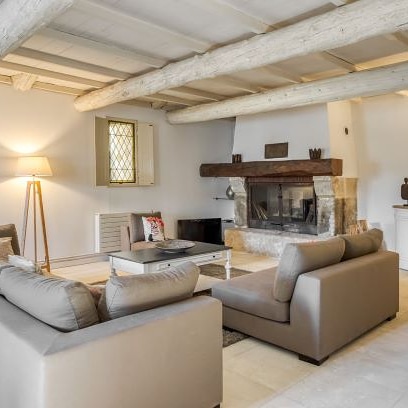 Un salon rustique et lumineux avec des poutres apparentes au plafond, deux canapés beiges, une table basse blanche, et une grande cheminée en pierre avec un manteau en bois.