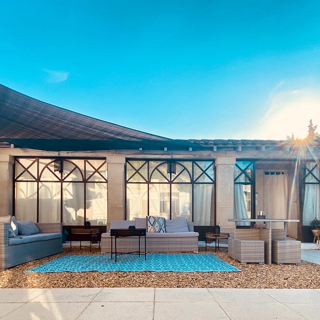 Une terrasse extérieure en gravier, aménagée avec des canapés et des fauteuils en rotin clair, une table basse, et une table à manger avec des poufs, sous une toile d'ombrage et devant une façade vitrée, le tout sous un ciel bleu.
